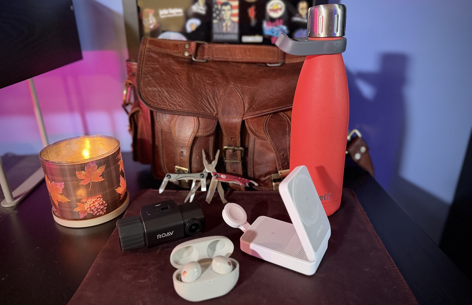 All five items from this article, the Gerber Dime, S'Well 17-ounce insulated bottle, ROAV Duo Dash Cam, Anker MagGo 3 and one folding travel charger, and the Sony WF-1000XM5 noise canceling wireless ear buds, on a leather mat. There's a burning candle and a leather bag in the background.