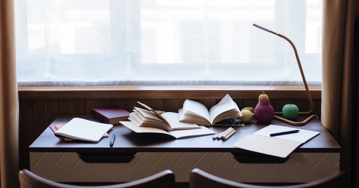 Image of a desk against a window. There is a city skyline outside of the window. On the desk there are multiple notebook, pens, and pencils.