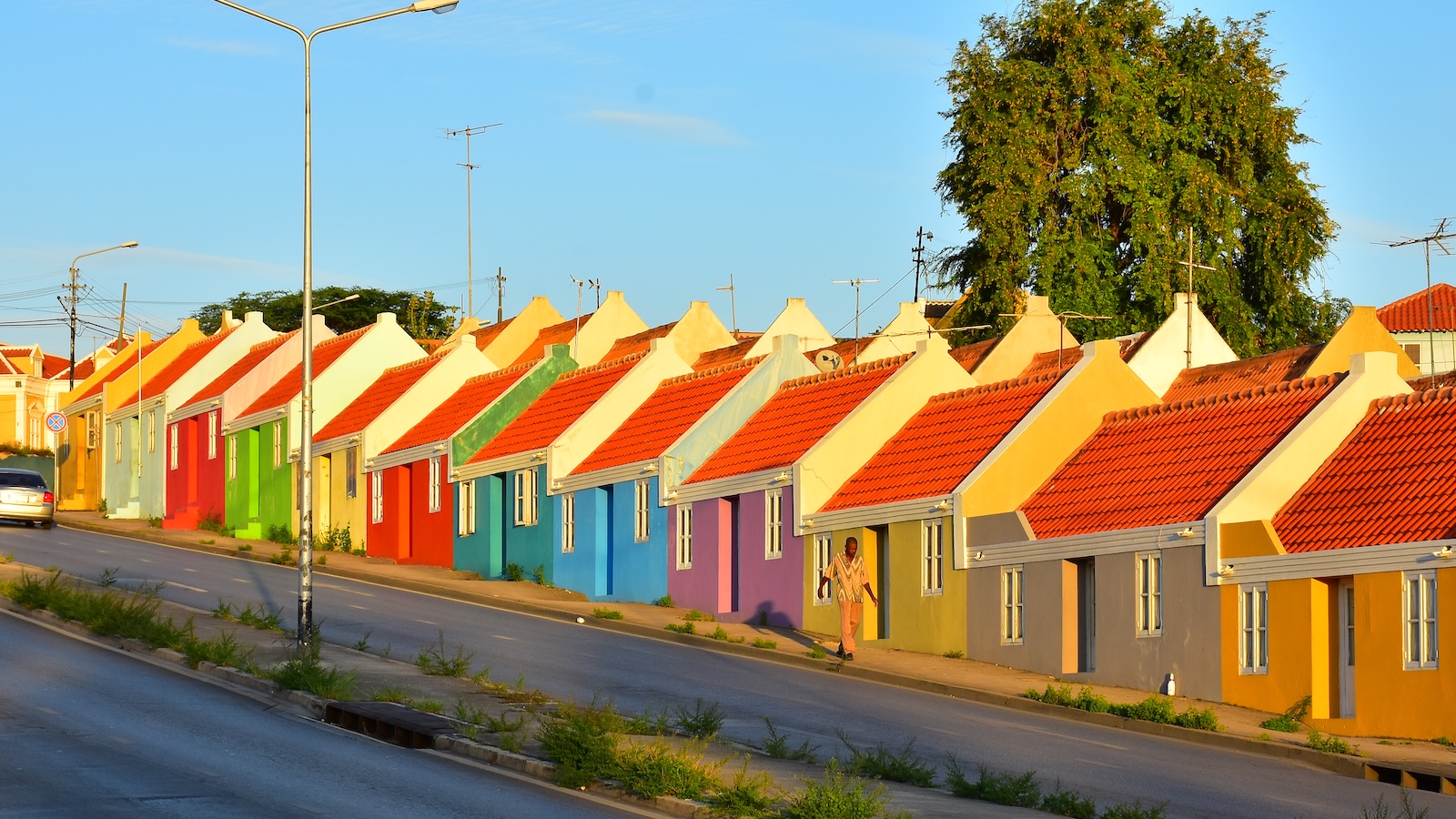 A row of colorful homes stretching uphill on a street in the Pietermaai neighborhood. They feature bright red tiled roofs. From, left to right they are painted gold, teal, red, green, yellow, red, turquoise, blue, purple, mustard yellow, purpl;e, and orange.