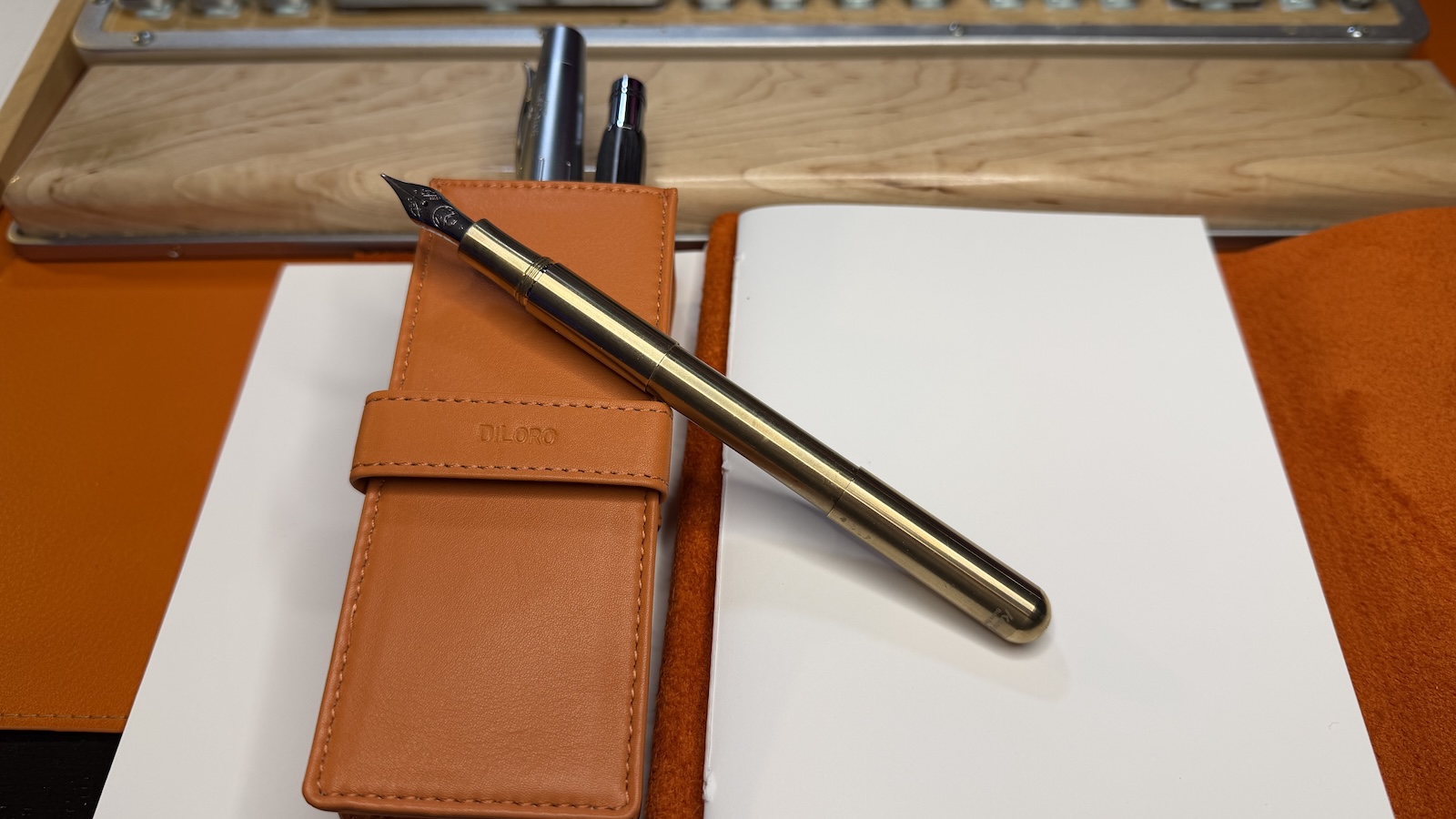 The Kaweco SUPRA Brass fountain pen open on a orange leather-bound note pad and an orange leather pen case.