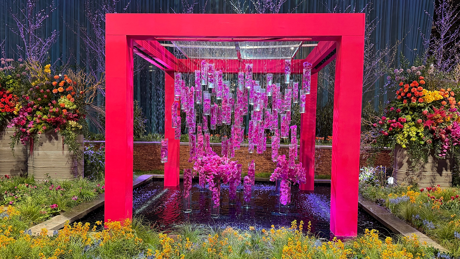 Philly-flower-show-4 A bright read pagoda spanning a pool. The pagoda has dangling fuschia floral fines. There are bright yellow and orange wild flower in the foreground, in front of the pool.