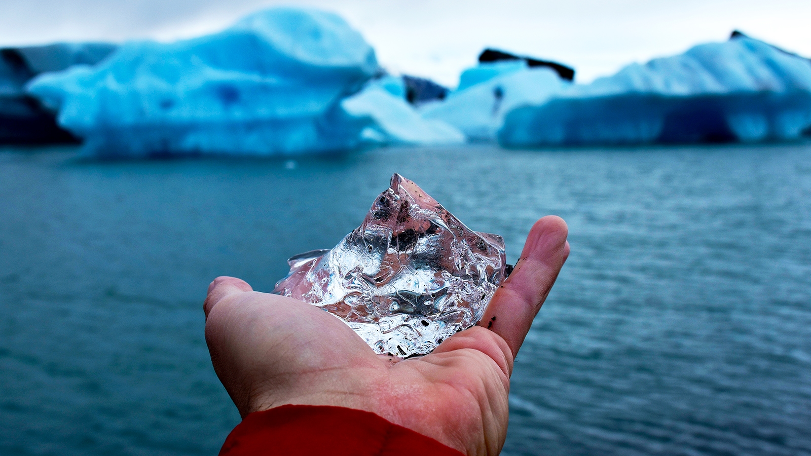 Reynisfjara A view of my hand holding a diamond-shaped piece of ice at Jökulsárlón Glacier Lagoon. Icebergs can be seen floating in the lagoon in the background.