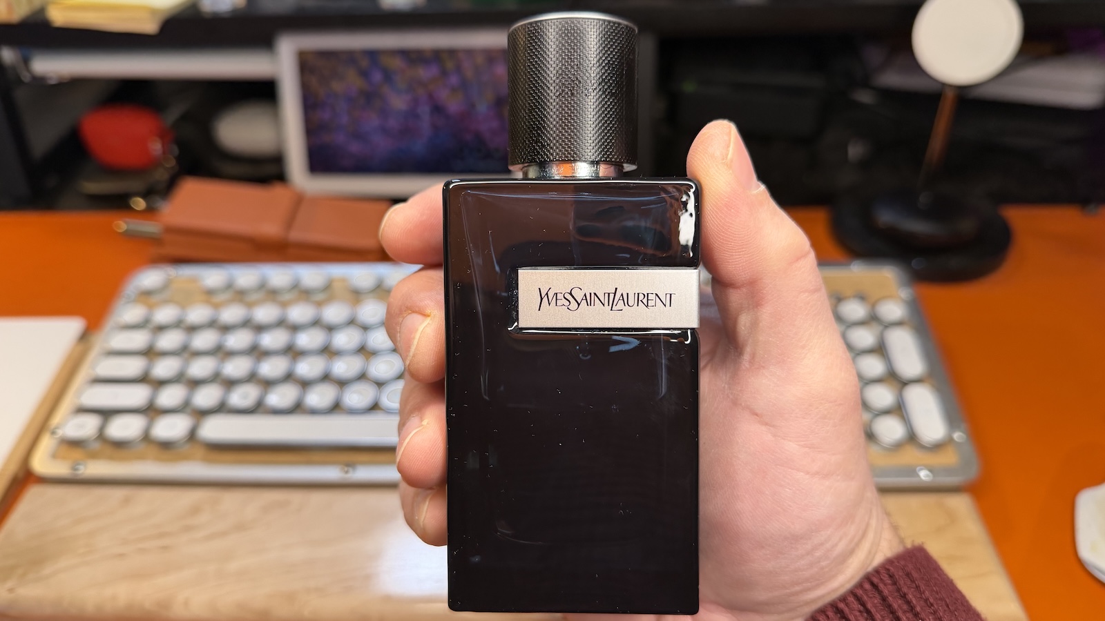 Yves-Saint-Laurent-Y My hand holding a bottle of Yves Saint Laurent Y, my desk top can be seen in the background, with my orange desk map and my maple wood mechanical keyboard.