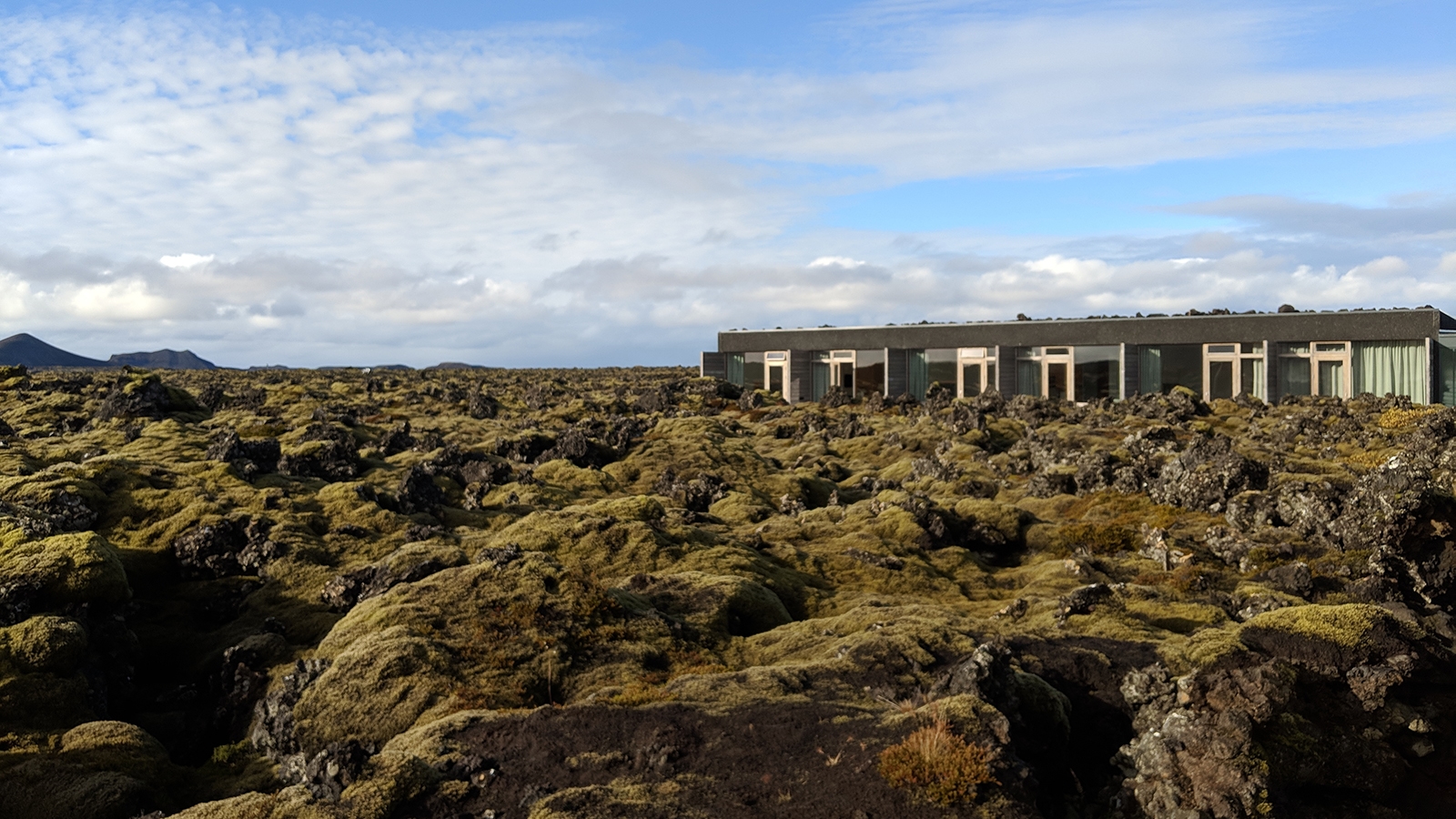 silica-hotel Hotel Silica viewed from the moss-covered lava boulder field, under a blue sky with fluffy white couds.