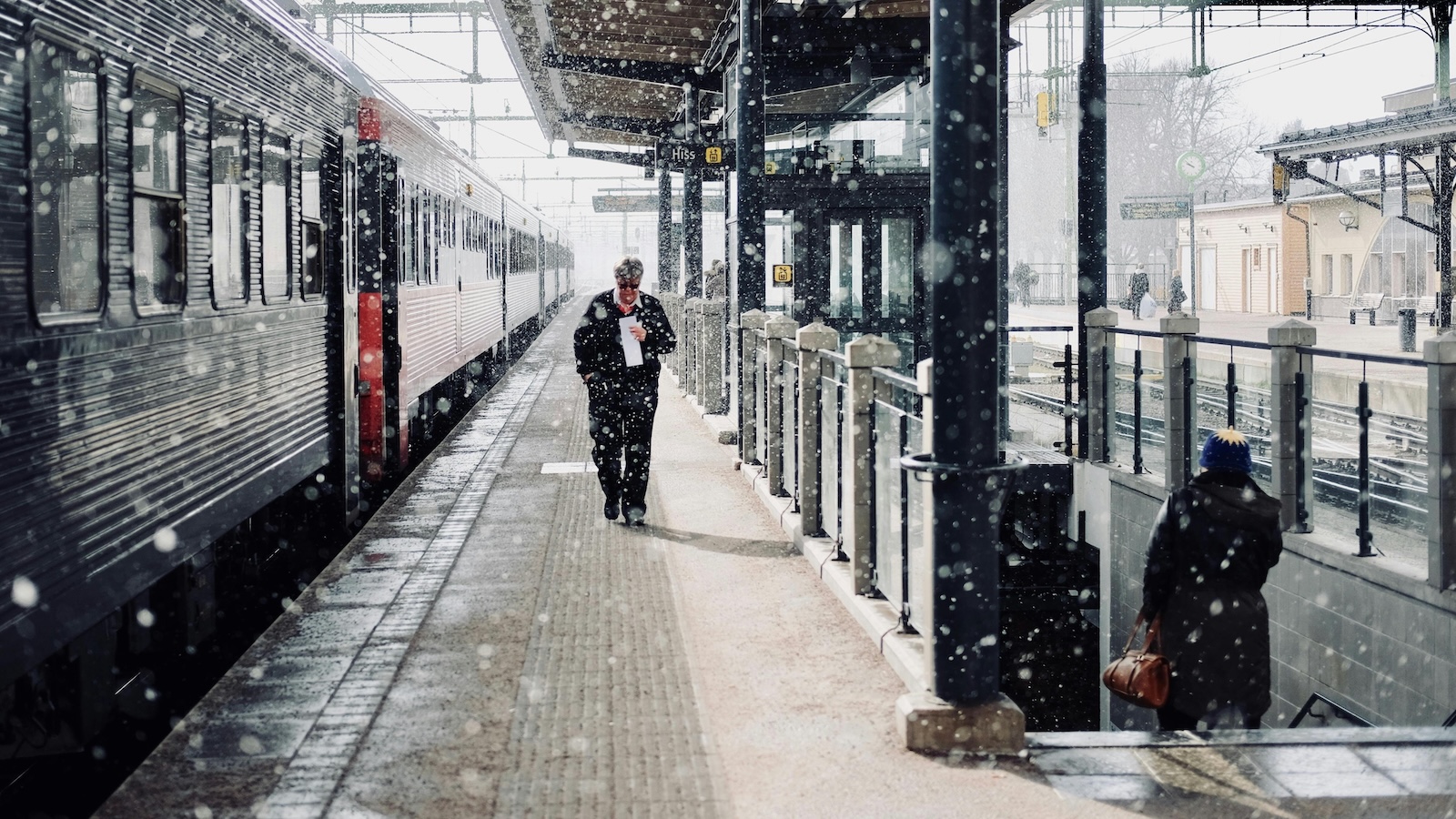 A man walking towards the camera on a train platform. A train is stopped at the platform, and it's snowing heavily.