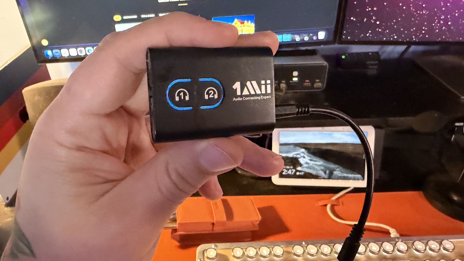 1Mii-bluetooth My hand holding the Mii Bluetooth 5.3 Transmitter Receiver, my office desk with my keyboard and displays can be seen in the background.
