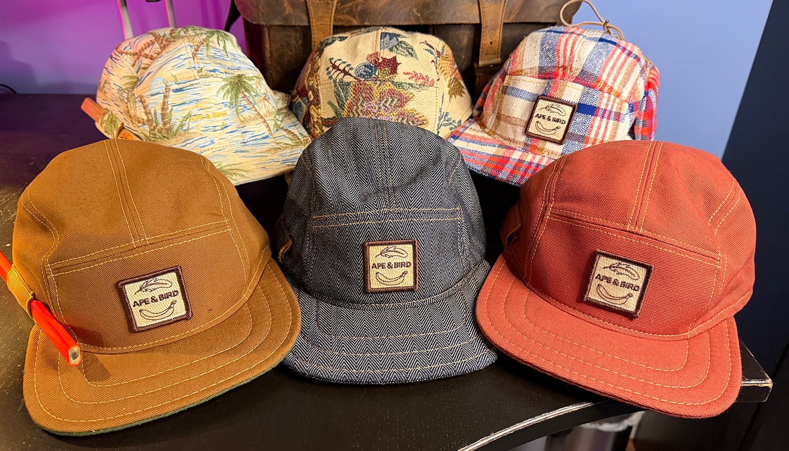 Six of my Ape & Bird Hats, in the back row, left to right, a pattern of a tropical beach scene with palm trees, sand, and waves, next Gramma's House, a burgandy, blue and green patten on a tan base, next is a bright red and blue plaid pattern with earflaps. In the front row, there's a solid camel colored hat, an indigo blue denim hat, and a a brick red solid hat.