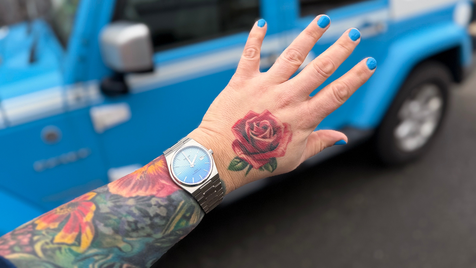 My hand and forearm covered in floral tattoos held out in front of my Chief Blue Jeep Wrangler. My nails are polished and the polish matches the blue of the Jeep. On my wrist is a Tissot PRX Automatic with a blue face.