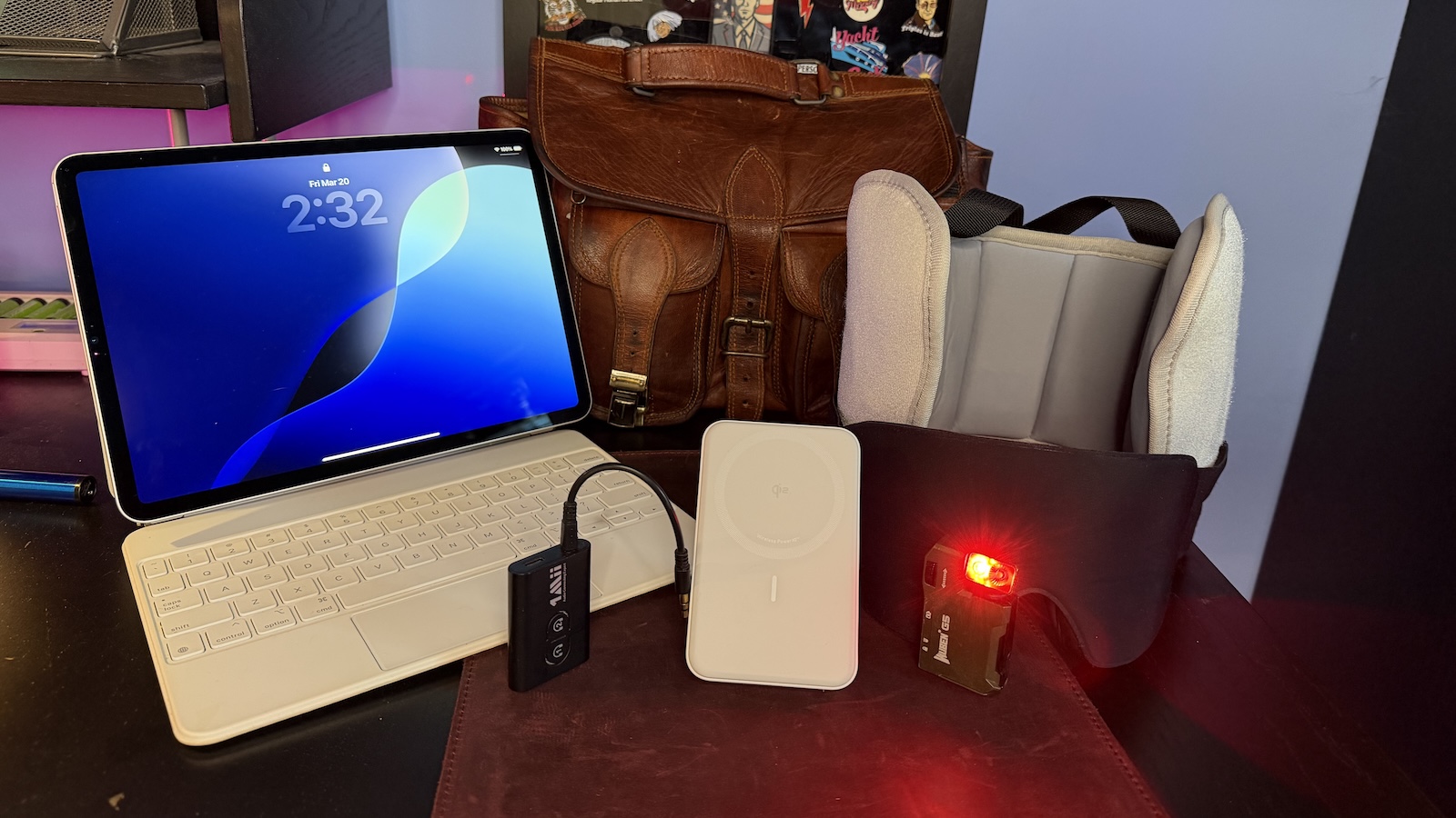 The five products featured in this article arranged on my desk. From left to right, there is the 11' iPad Pro, 1Mii Bluetooth 5.3, Transmitter Receiver, Anker MagGo 10K Power Bank, the WUBEN G5 Rechargeable EDC Flashlight, and the SARISUN Travel Pillow. In the background is a leather messenger bag and an enamel pin case mounted on the wall.