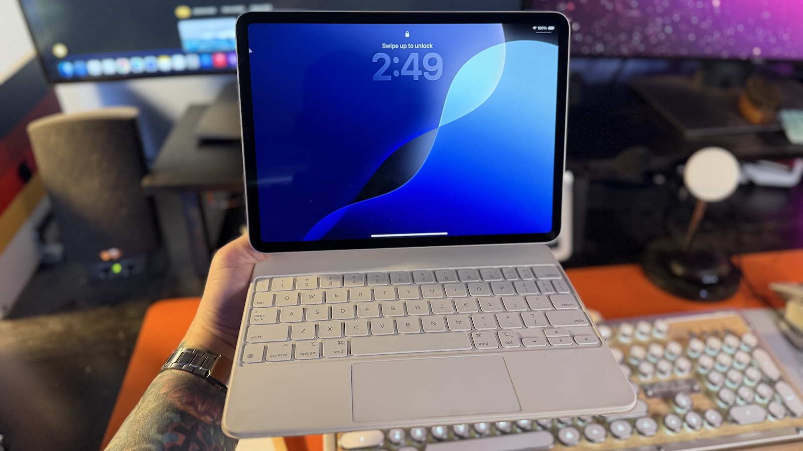 My arm holding an 11-inch iPad Pro with Magic Keyboard, my office desk with my keyboard and displays can be seen in the background.