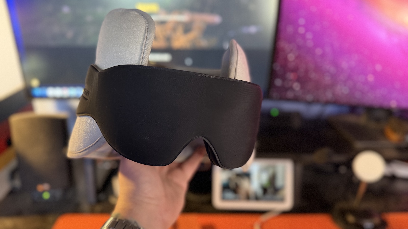 My hand holding the SARISUN Travel Pillow, my office desk with my displays can be seen in the background.