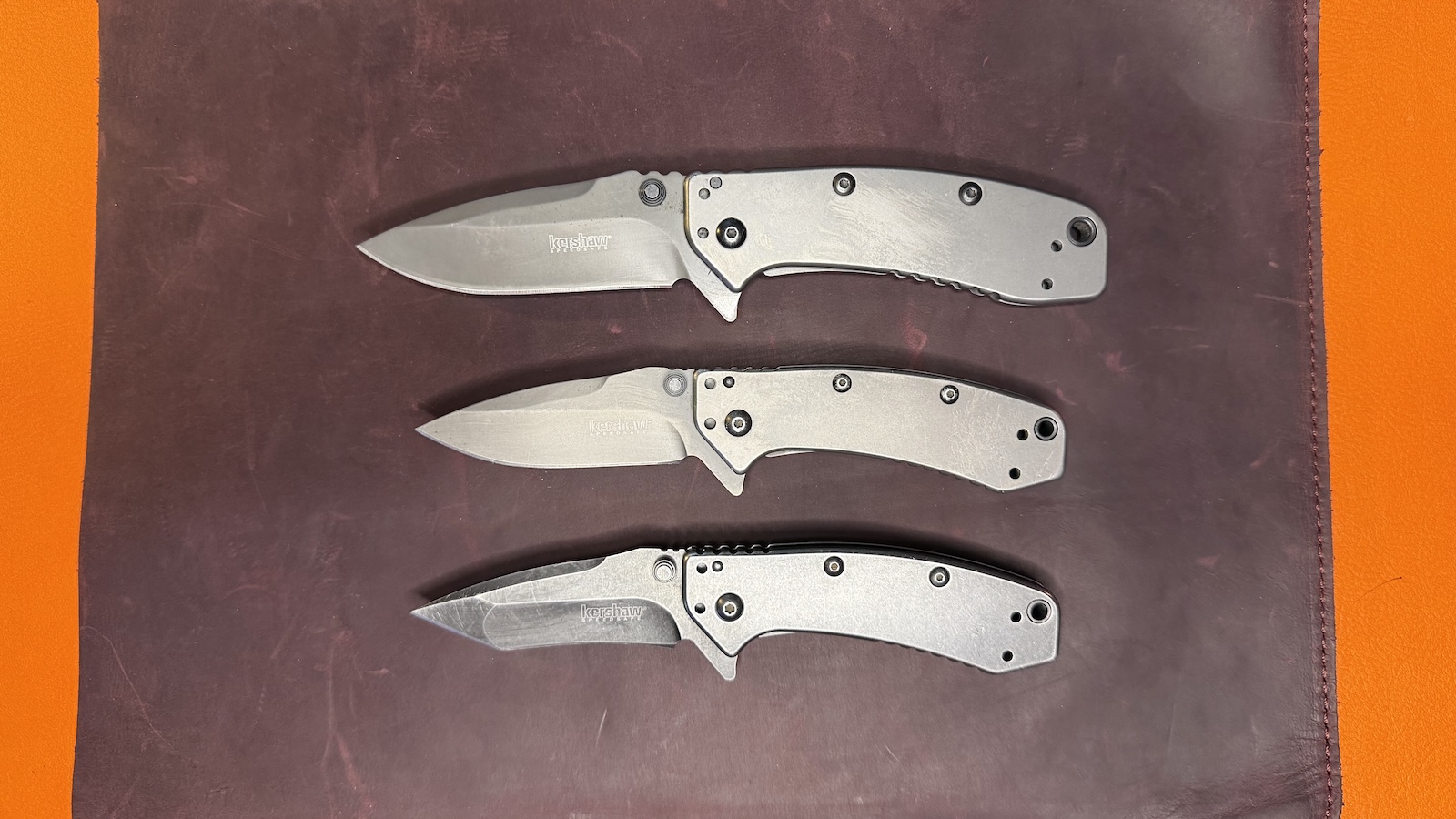 Kershaw-Cryo Three Kershaw Cryo pocket knives are sitting on brown leather. From top to bottom: The Cryo II drop point, The Original Cryo drop point, and the Cryo tanto blade.