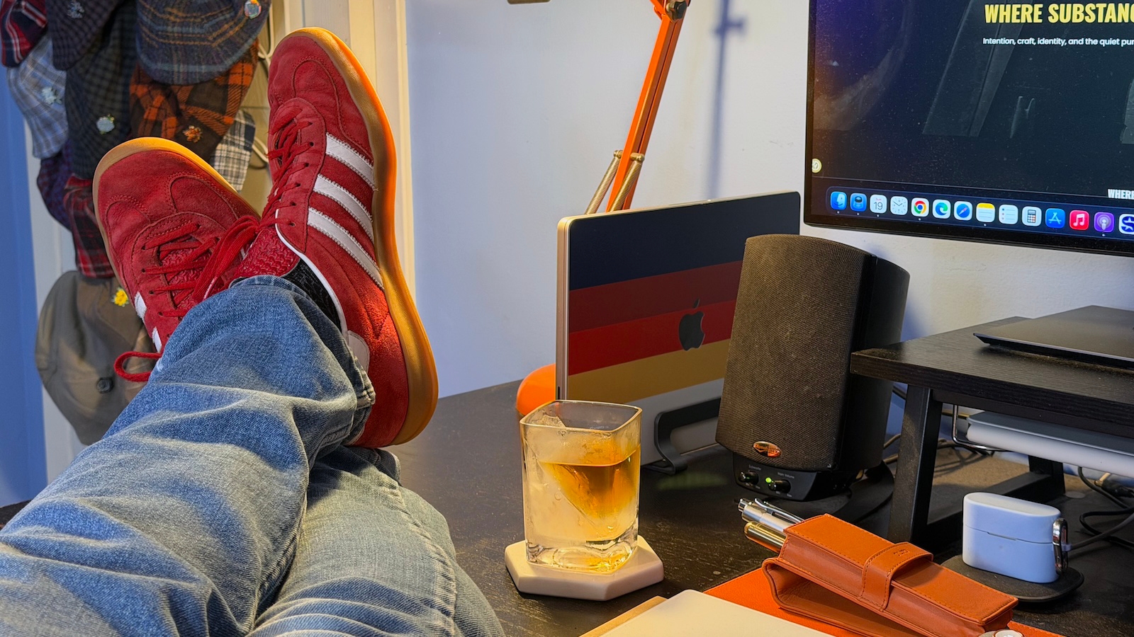 My feet up on my desk, wearing blue jeans and a pair of Adidas Gazelle Indoors in Scarlett with cloud white stripes. On my desk you can see my MacBook Pro, a Klipsch speaker, my fountain pen case, and an ice wedge glass containing scotch.