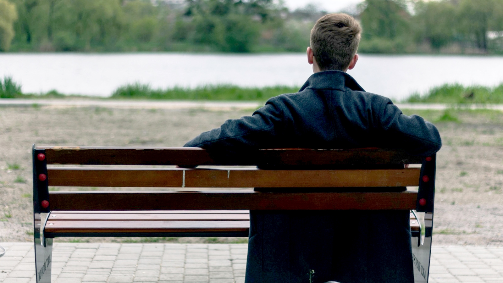 A man sitting on a park bench staring at a lake, the image is shot from behind.
