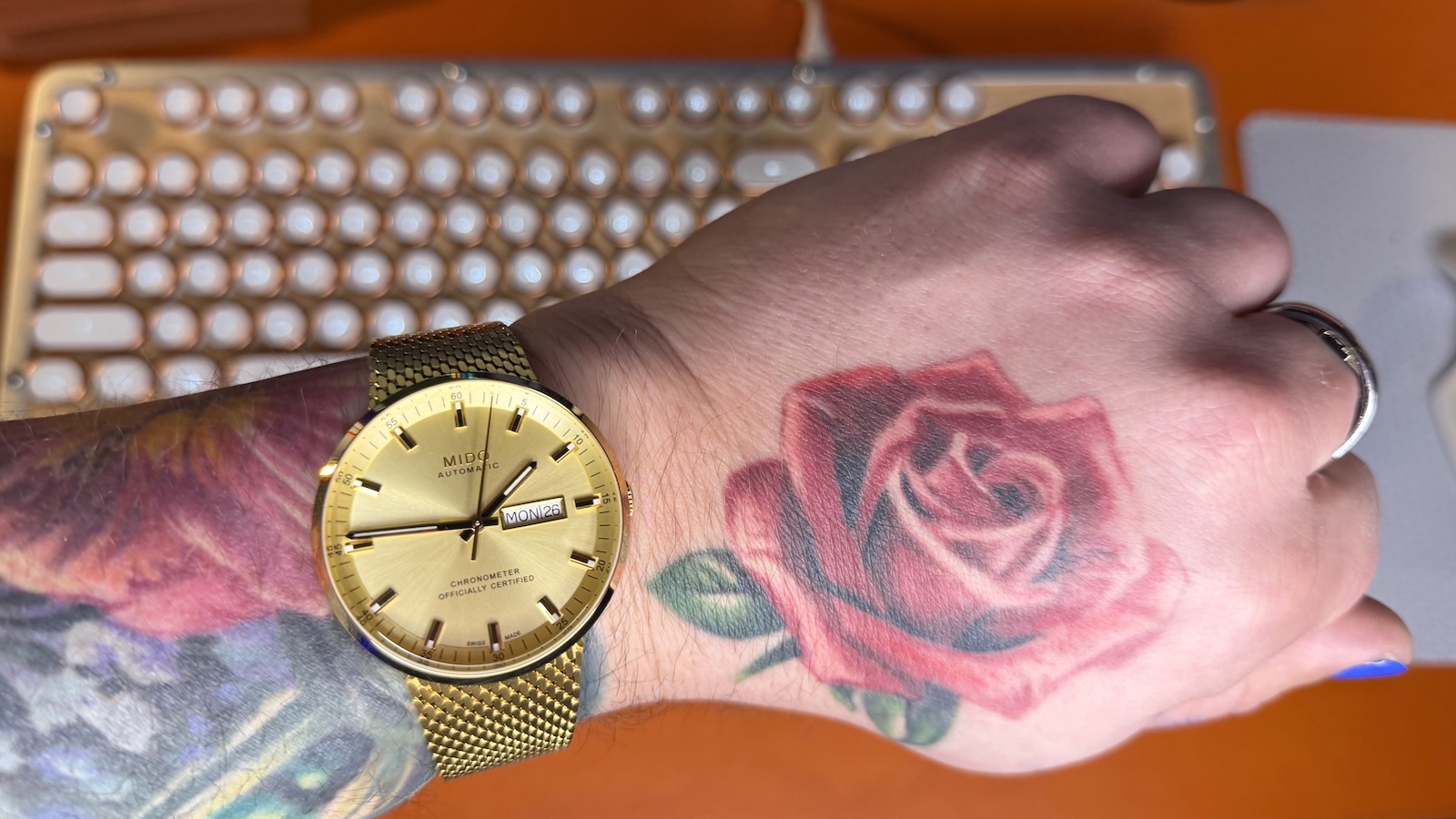 The Mido Commander Ic&ocirc;ne on my wrist, my forearm is heavily tattooed and there's a rose tattoo on the back of my hand. An illuminated AZIO Retro Classic keyboard in light wood can be seen blurred in the background.