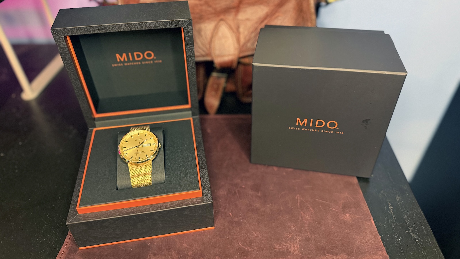 The Mido Commander Ic&ocirc;ne sitting in its box on a leather mat. A leather messenger bag can be seen blurred in the background.