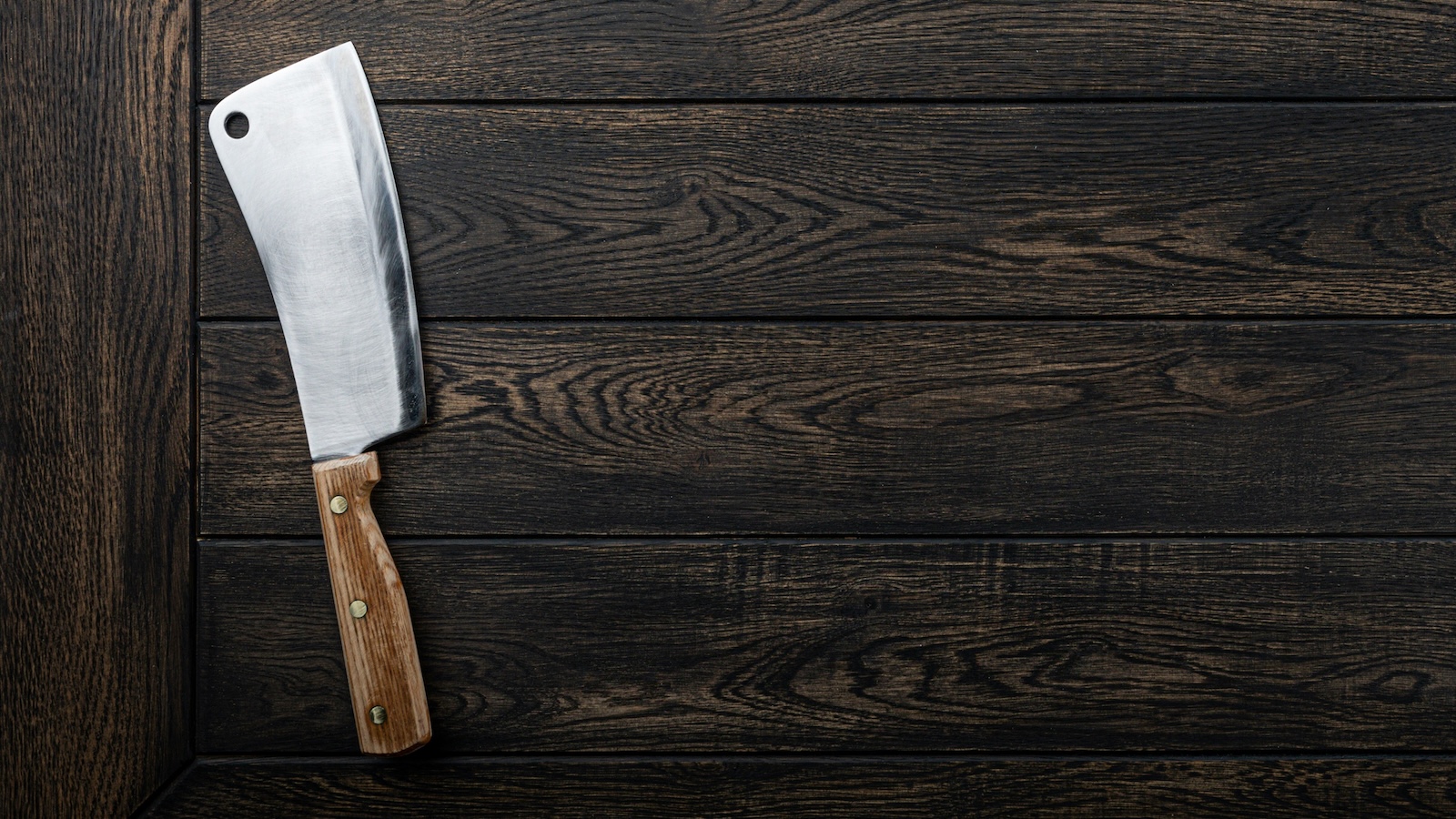 A meat cleaver with a light wooden handle sitting on the dark wooden tabletop.