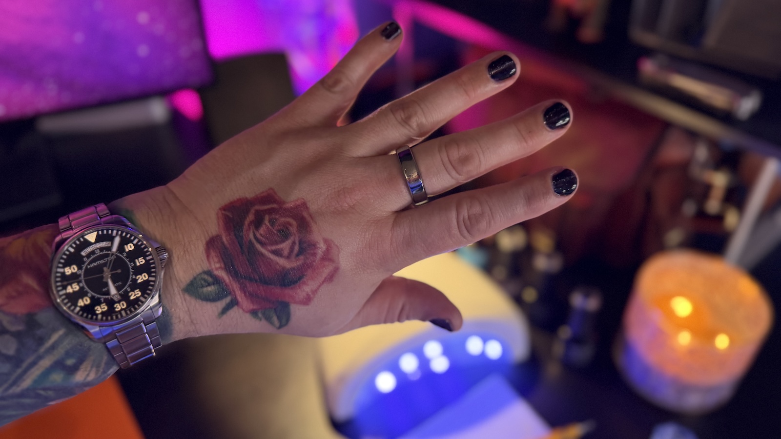 My left hand and forearm, my nails are painted dark purple with silver cat-eye effects, my hand has a rose tattoo, and on my wrist I'm wearing a Hamilton Pilot Day Date watch. My forearm is heavily tattooed. Blurred in the background, you can see an array of nail polishing tools, a UV lamp, and bottles of polish.