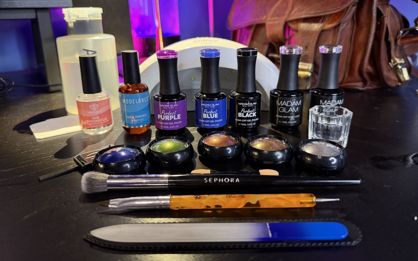 painting-nails-gel-polish An array of gel polish necessities, including a pump alcohol dispenser, lint-free wipes, a SUNUV Sun 1 lamp, modelones nail dehydrator and primer, Madam Glam Perfect Purple, Perfect Blue, and Perfect Black, Madam Glam basecoat and topcoat, a small class cup for holding alcohol, a magnet for cat-eye effects, chrome powders in blue, green, orange, gold, and silver, and eye shadow applicator and brush for applying chrome powder, and No 2. polish brush, and a glass nail file.