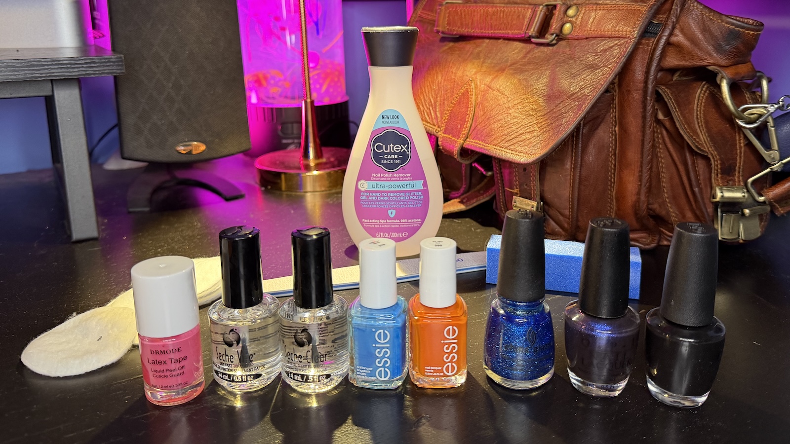 painting-nails-standard-polish An array of standard nail polishing necessities, including a botle of Cutex nail polish remover, cutton pads, an emery board file, and sponge buffer, liquid latex tape, Seche Vite fast drying basecoat and topcoat, Essie polishes in blue and orange, and polishes by other brands in blue glitter, purple, glitter, and black.
