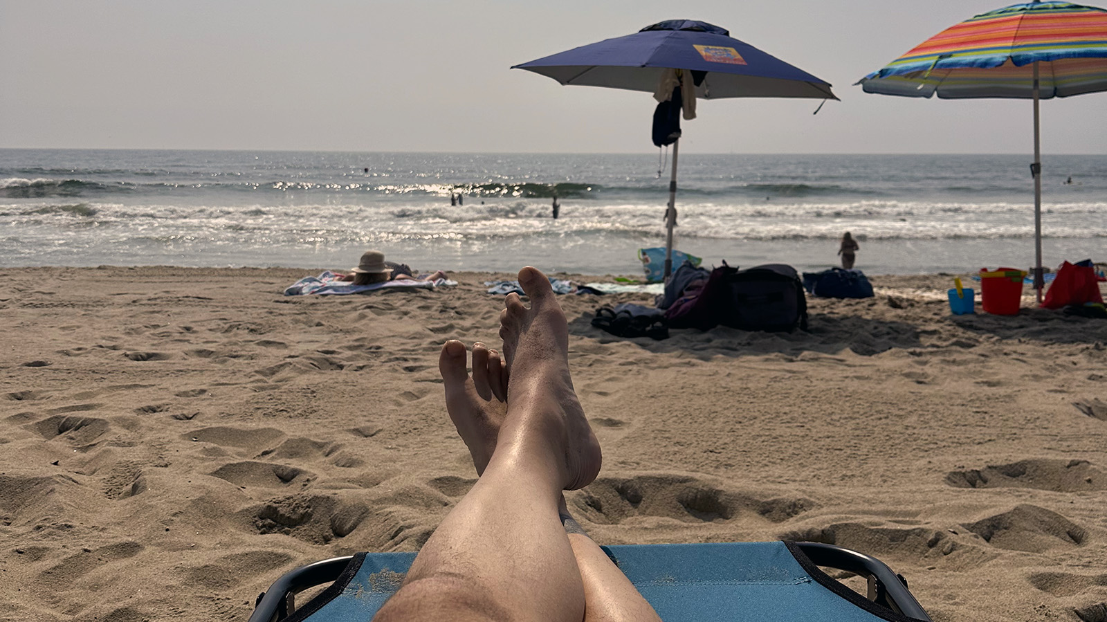 A beachgoer rests on a low chair with sandy feet pointed toward the ocean, with umbrellas, shoreline, and waves in the background on a sunny beach day.