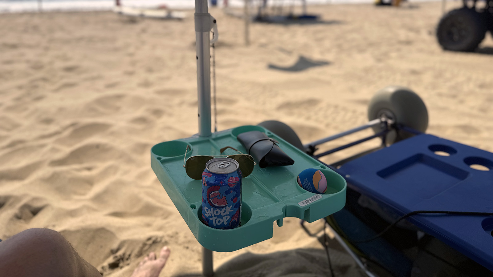 beachbub-table BeachBub umbrella table holding a drink, sunglasses, and accessories beside a beach cart on the sand.