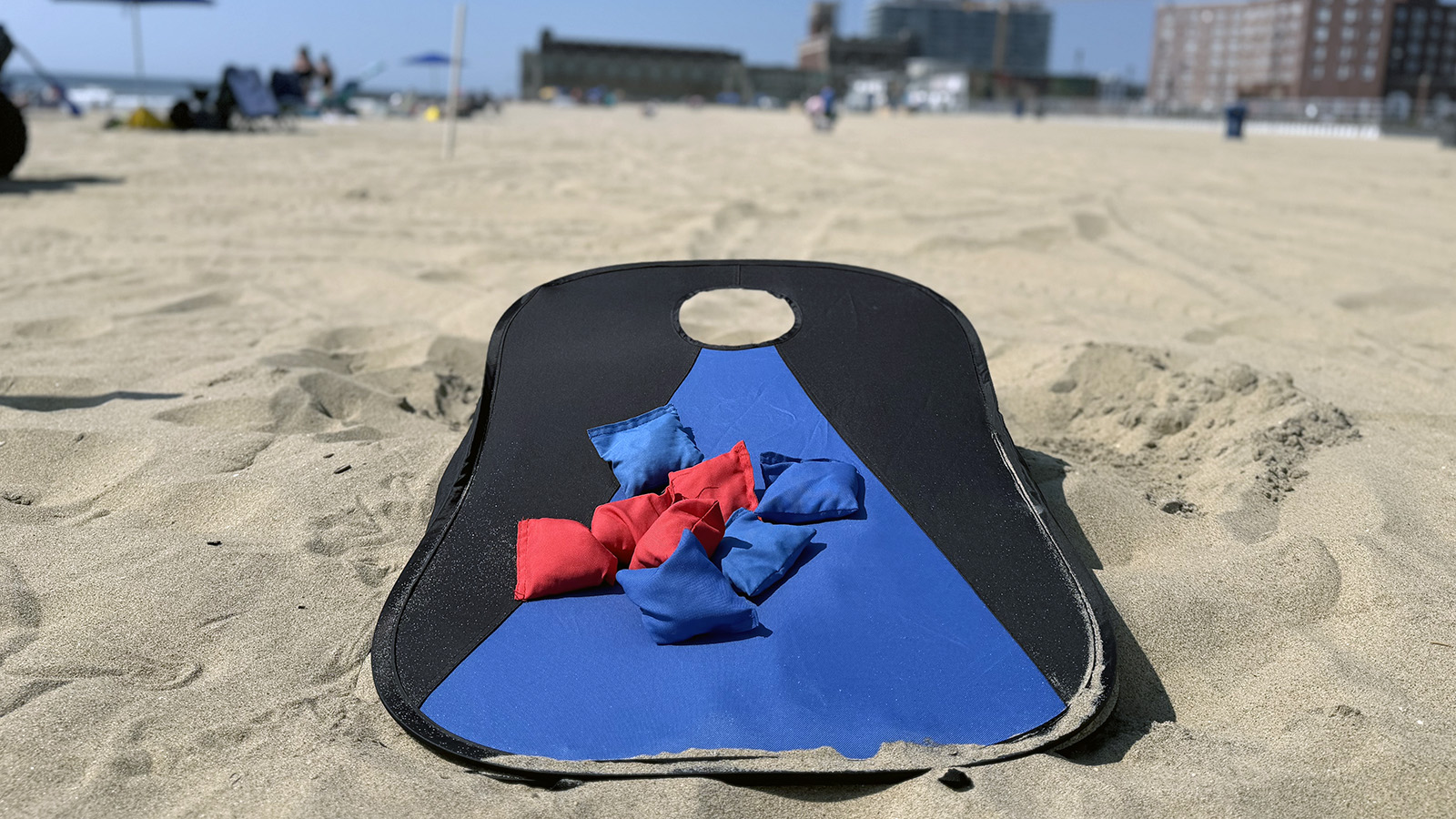 cornhole Portable blue and black cornhole board with red and blue beanbags set up on the beach.