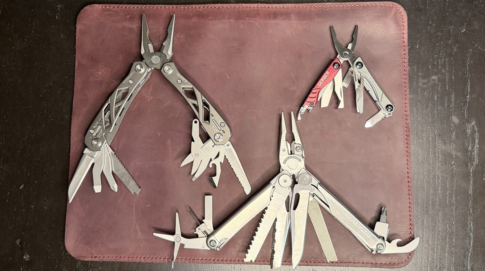 The Gerber Suspension, The Gerber Dime, and The Leatherman Wave Plus on a leather mat placed on a black wood background.