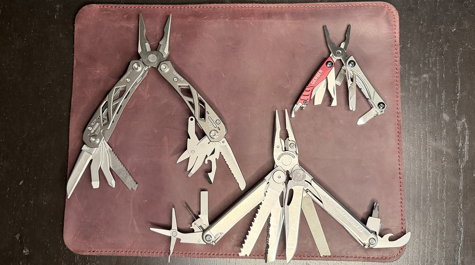 The Gerber Suspension, The Gerber Dime, and The Leatherman Wave Plus on a leather mat placed on a black wood background.