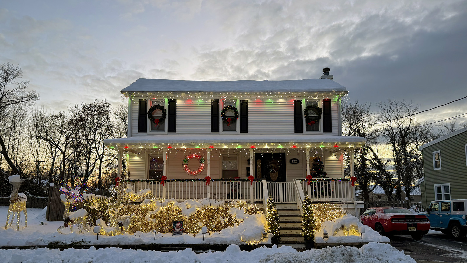 porch-christmas Snow-covered farmhouse with a full-width porch decorated with Christmas lights, wreaths, garland, and illuminated holiday figures.
