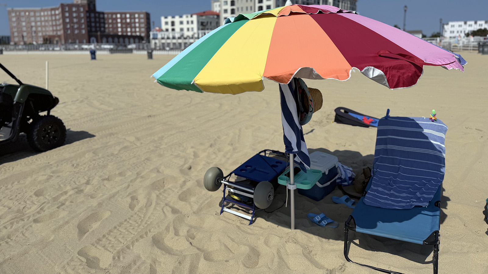 umbrella Large rainbow beach umbrella shading a chair, cooler, table, and beach cart on the sand.