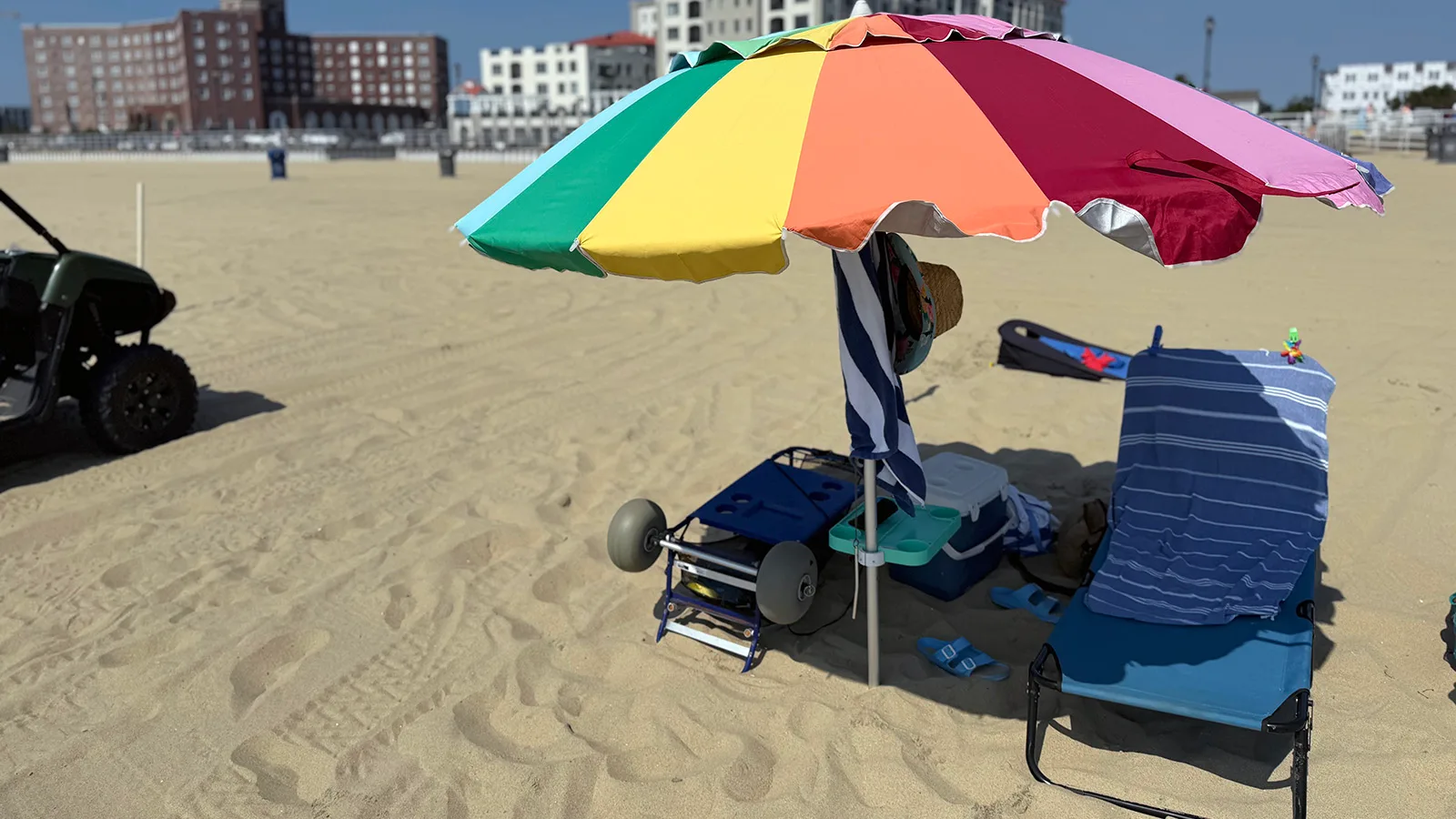 Large rainbow beach umbrella shading a chair, cooler, table, and beach cart on the sand.