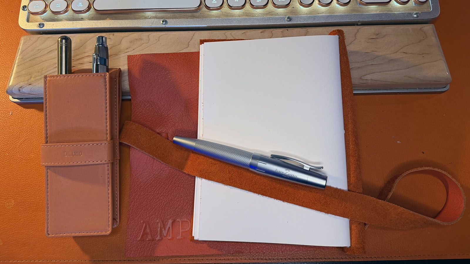 Silver Faber-Castell e-motion fountain pen resting across an open leather notebook, beside a brown leather pen case containing two other pens, on an orange desk.