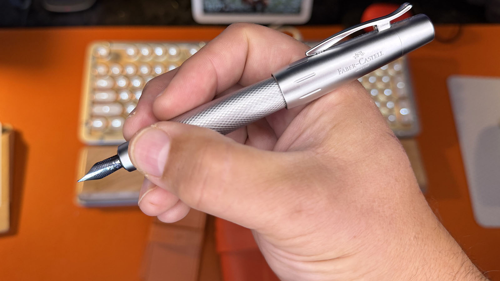 Close-up of a hand holding an uncapped Faber-Castell e-motion fountain pen in writing position, displaying its exposed metal nib, textured silver barrel and posted cap.