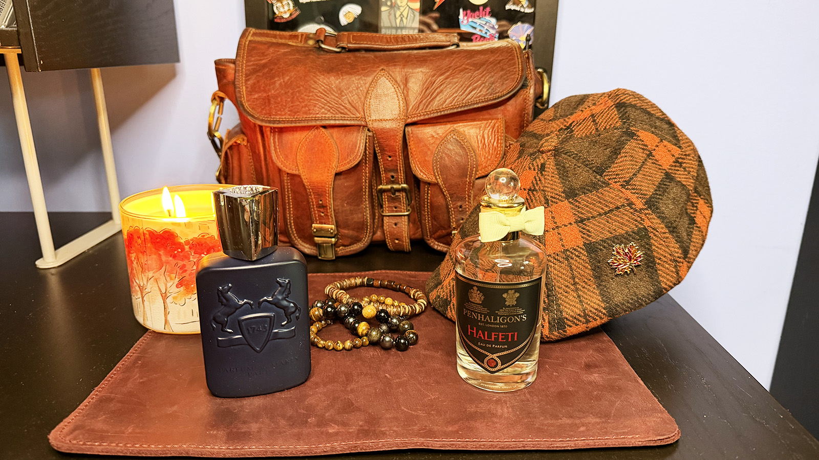 Parfums de Marly Layton and Penhaligon’s Halfeti fragrance bottles displayed with a lit candle, leather satchel, beaded bracelets, and an orange-and-brown plaid cap.
