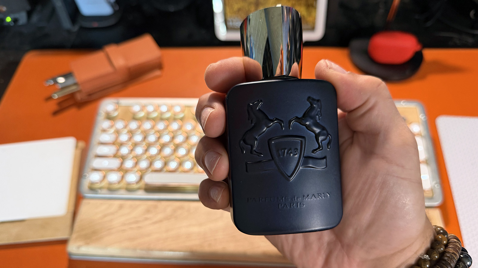 Hand holding a dark blue Parfums de Marly Layton fragrance bottle in front of an orange desk with a keyboard softly blurred in the background.