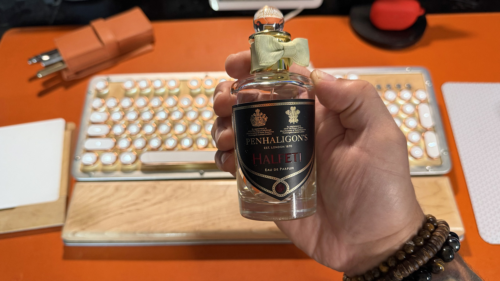 Hand holding a clear Penhaligon’s Halfeti fragrance bottle in front of an orange desk with a keyboard softly blurred in the background.