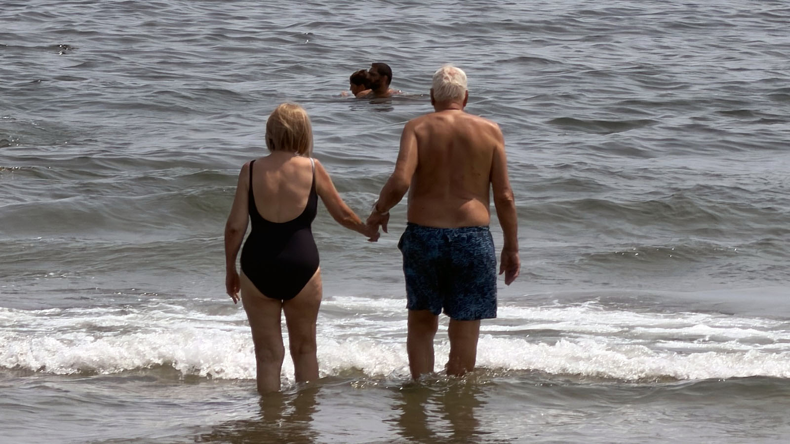 An older couple (my parents) holding hands as they walk into the ocean at the beach.