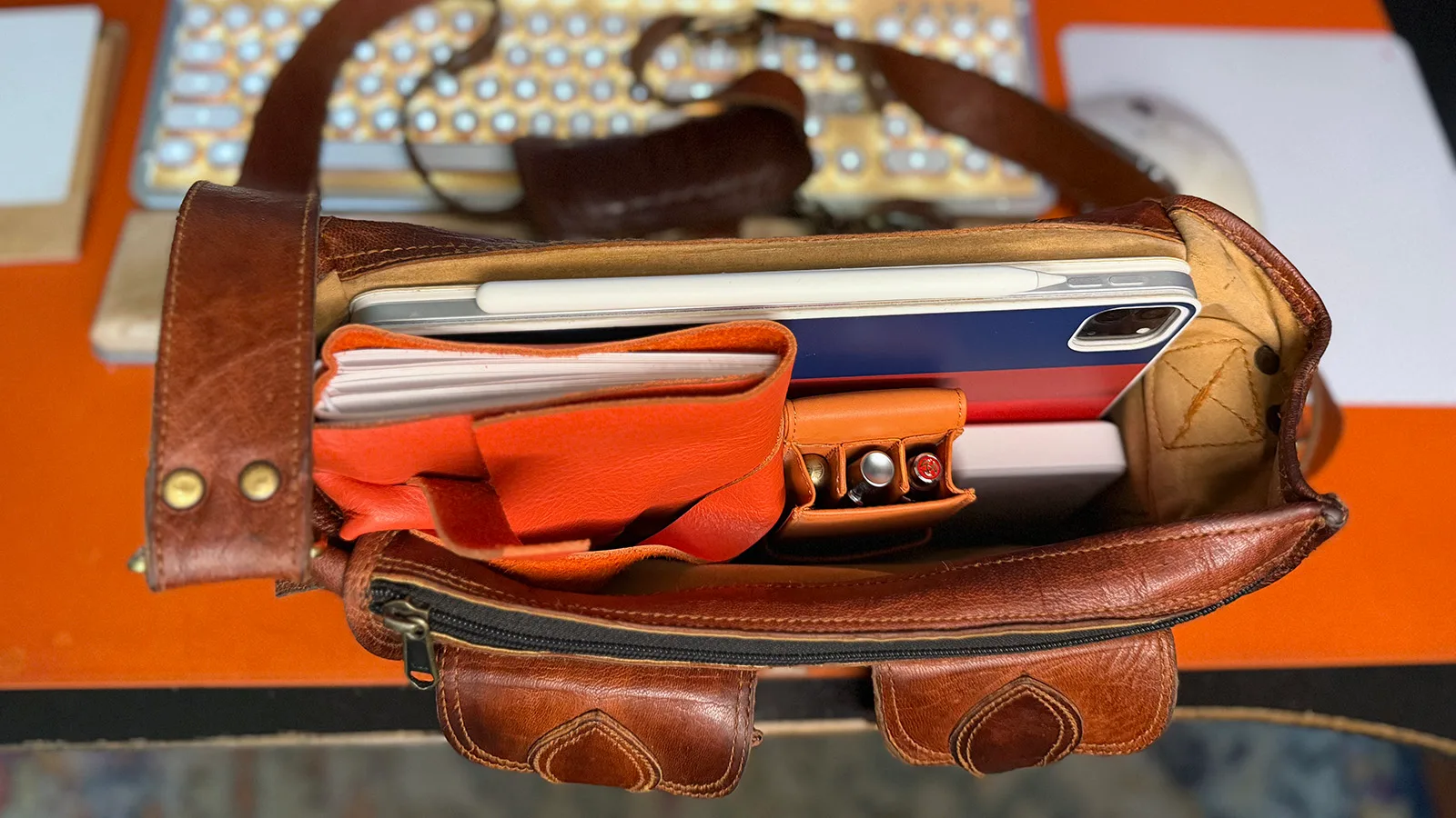 my-bag-contents Open leather crossbody bag packed with an iPad, notebook, pen case, and other travel essentials arranged inside.