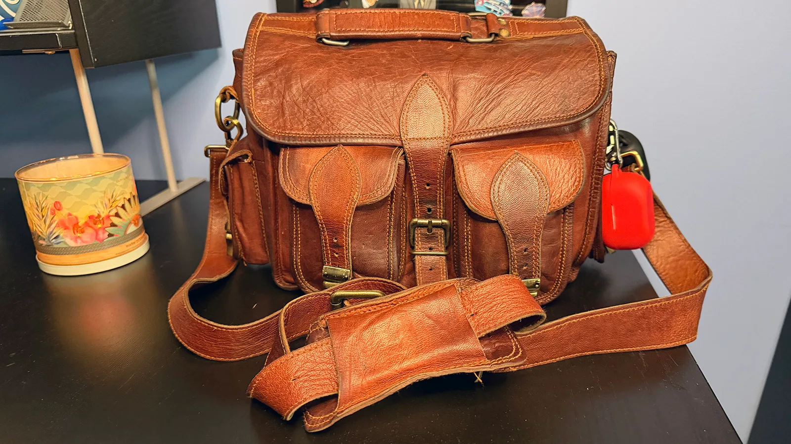 my-personal-item Worn brown leather crossbody travel bag with front pockets, brass hardware, shoulder strap, and clipped earbud case.