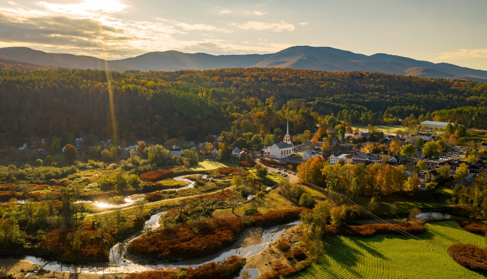Aerial view of Stowe village surrounded by autumn foliage, with a white church steeple, winding river, and mountains in the distance.